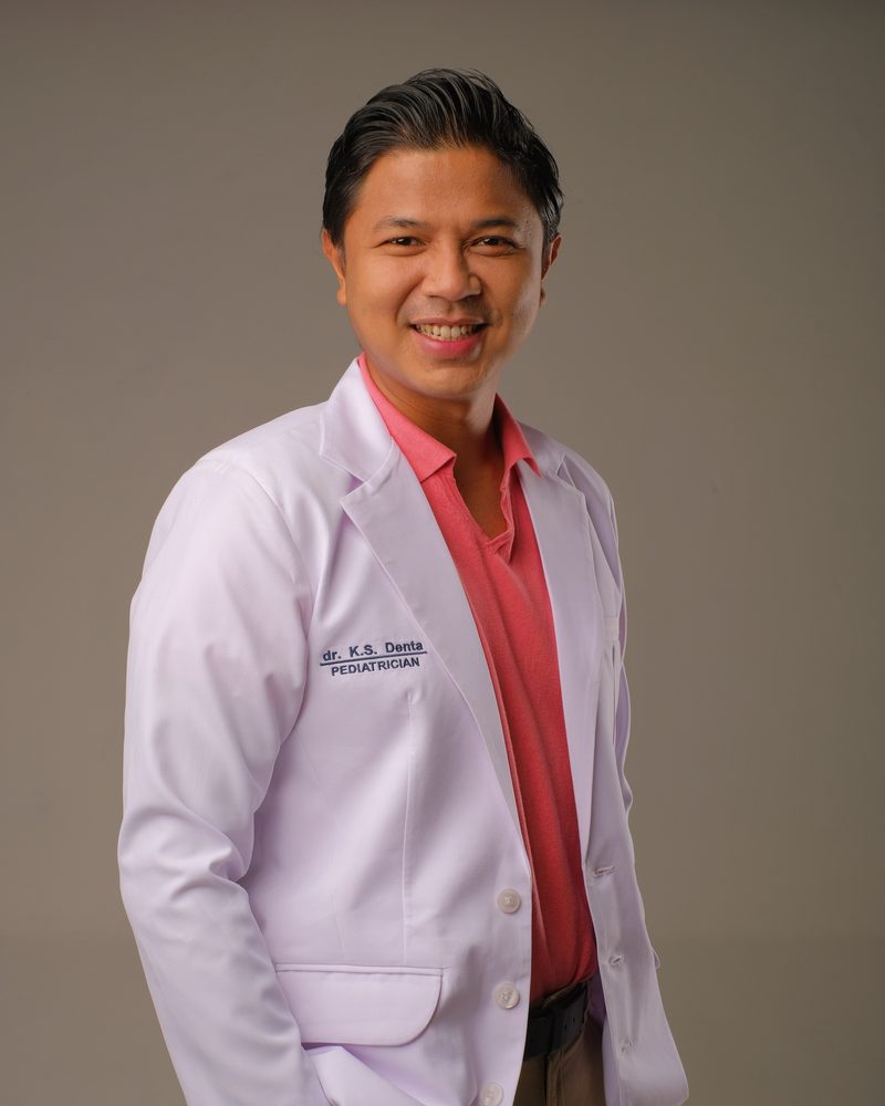 dr. Kurniawan Satria Denta, dokter spesialis anak, tersenyum mengenakan jas dokter putih bertuliskan dr. K.S. Denta Pediatrician dan kemeja merah muda, berlatar abu-abu